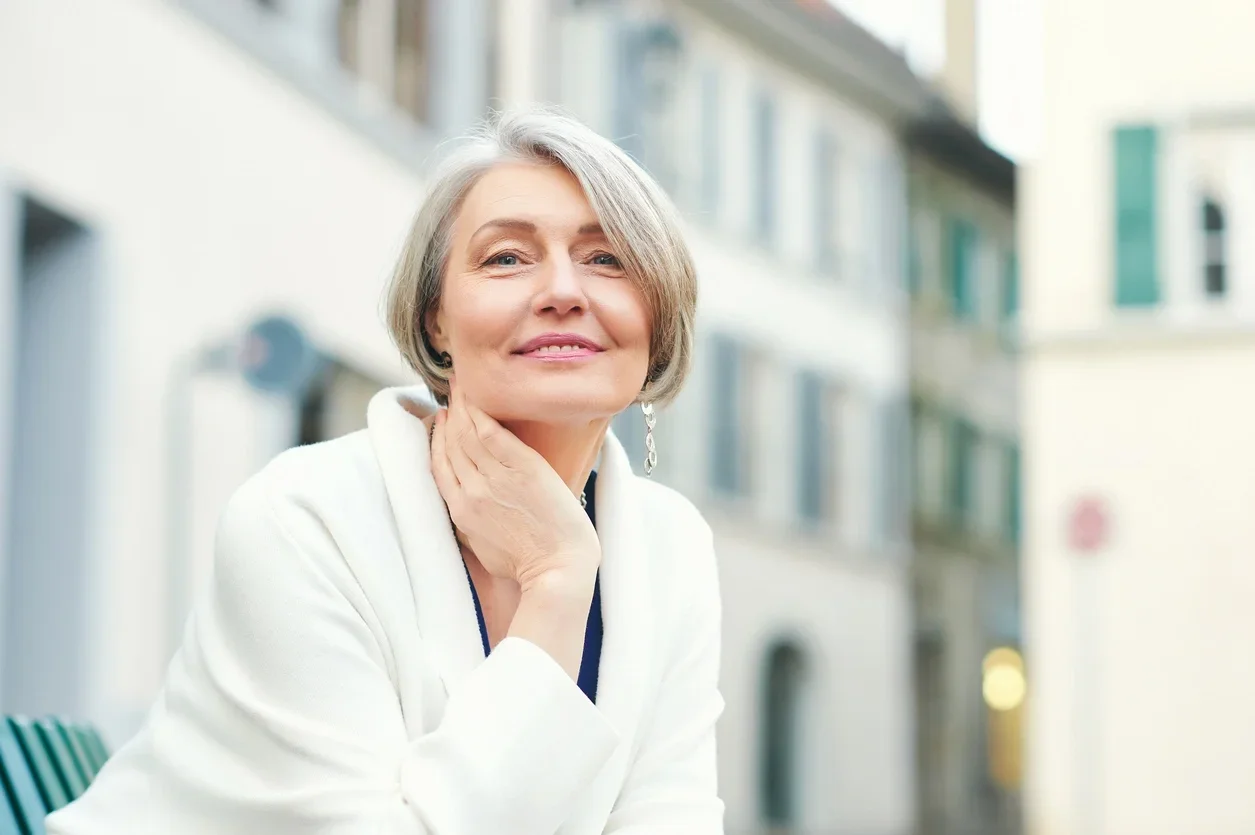 Retrato ao ar livre da linda e elegante mulher de meia-idade de 55 a 60 anos, vestindo jaqueta branca, sentada no banco do lado de fora com corte de cabelo pixie
